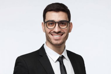 Yatay headshot gençlerin Avrupa beyaz adam resmi kıyafetler gözlüklerle bakıyor ve olumlu, gülümseyen gri arka plan üzerinde izole duygu onun iş dünyasında başarı hakkında kendinden emin