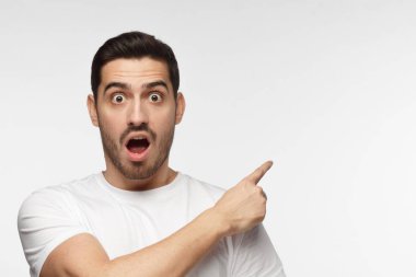 Horizontal photo of young Caucasian man pictured isolated on gray background dressed in white T-shirt looking at camera with mouth and eyes open with surprise, pointing right, copyspace for advert clipart