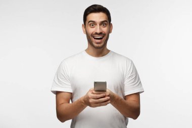 Yatay fotoğraf genç yakışıklı Avrupa adamın gri arka plan ayakta beyaz T-shirt holding cep elinde izole gözleri sevinçle sanki ödül kazandığı gülüyor sürpriz ile yuvarlak.