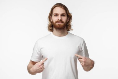Stüdyo portre genç yakışıklı erkek ile beyaz arka plan beyaz boş T-shirt giymiş izole uzun sarı saçlar ve her iki ellerini, pozitif, boşaltmak için reklam ile bunu işaret eden sakallı