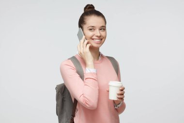 Genç yakışıklı genç kızın resimde yatay atış kahve fincanı ile elinde duran gri arka plan üzerinde izole ve gülümseyen, duygu sakin ve rahat smartphone üzerinde konuşurken sırtına sırt çantası