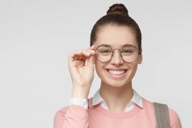 Avrupa beyaz bir kadın resimde genç gri arka plan üzerinde izole closeup portresi, saçlarını hairbun, holding kenarında büyük bağlı yuvarlak gözlük mutlu karşılama gülümseme gösterilen parmaklı