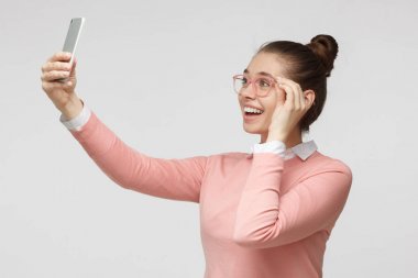 Horizontal photo of young European female in casual clothes and glasses isolated on grey background taking selfie picture with cellphone smiling happily as if expecting to get likes in social networks clipart