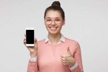 Pembe kazak ve boş smartphone ekran gülümseme, boşaltmak için reklam ile gösterilen gri arka plan üzerinde izole gözlük, stüdyo portre genç yakışıklı Avrupa kadın giyinmiş