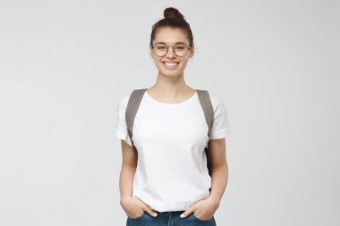 Genç yakışıklı genç kızın içinde rahat ve olumlu, yürümeye istekli hissediyor gözlük ve gri sırt çantası ve kot pantolon, beyaz casual T-shirt, gri arka plan üzerinde izole kapalı resim