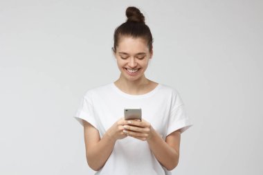 Yatay portre genç olumlu Avrupa beyaz beyaz T-shirt giyen, gülümseme ile akıllı telefon ekranına bakarak, ileti değiş tokuşu veya web sayfalarında gezinmeye gri arka plan üzerinde izole kadın