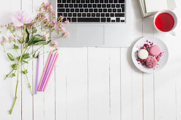 Izolované ploché zobrazení pracovního stolu malované bílé s kovovou laptop obklopen květinami, šálek čaje a deska s macarons, spoustu pastelek, ležící vedle připravené pro kreativní kreslení — Stock fotografie