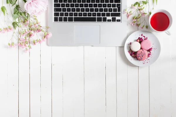 Isolated flat top view of wooden desk painted white with gray open laptop, black keyboard, light pink flowers, white saucer with pink and white macarons and cup of tea, ready for joyful freelance work — Stock Photo, Image