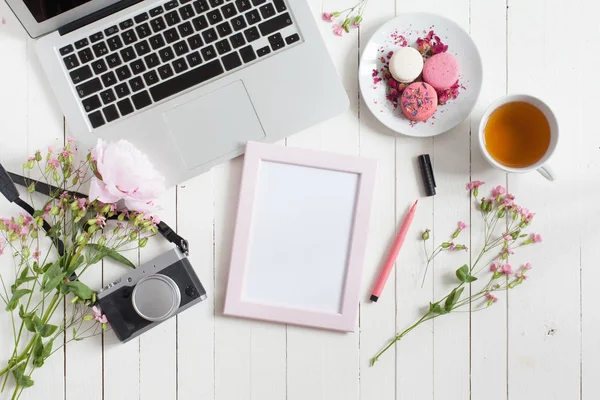 Tampilan atas rata meja kerja putih dengan laptop logam abu-abu dengan keyboard hitam, ditutupi dengan bunga merah muda, dengan kamera, bingkai foto merah muda dan pena ujung merasa untuk sketsa, makaron dan cangkir teh — Stok Foto