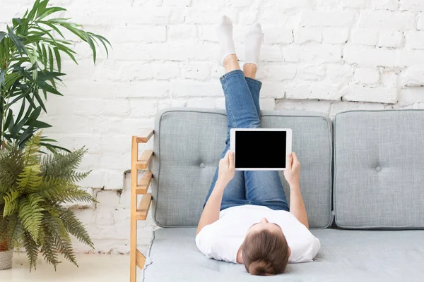 Horizontální obrázek Evropská dívka na obrázku uvnitř ležící na šedou pohovku s nohama pozdvihl hospodářství tabletový počítač s prázdnou obrazovkou, copyspace za své zboží nebo služby — Stock fotografie