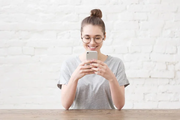 Estudio de la joven hermosa mujer caucásica sentada en la mesa de madera contra la pared de ladrillo blanco vestida con una camiseta casual, con gafas redondas, mensajes de texto o navegación con ayuda de un teléfono inteligente —  Fotos de Stock