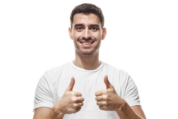 Closeup picture of young handsome European Caucasian male isolated on white background dressed in white T-shirt, showing thumbs up and looking positive, recommending product or service of quality