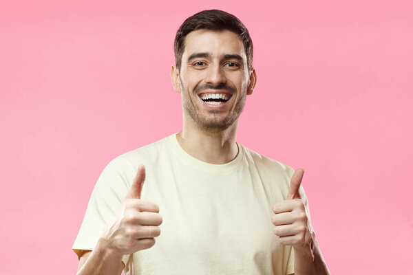 Indoor picture of young European male isolated on pink background wearing casual T-shirt smiling happily and showing thumbs up as if recommending service or product of good quality he has tried
