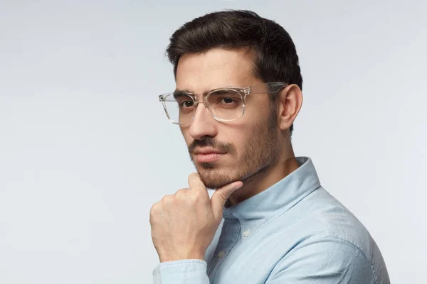 Zbliżenie Studio młodych europejskich kaukaski mężczyzna na zdjęciu na białym tle na szarym tle sobie fryzury krótkie i plastikowe okulary przezroczysty, patrząc na bok i drapanie jego podbródek myśląc — Zdjęcie stockowe