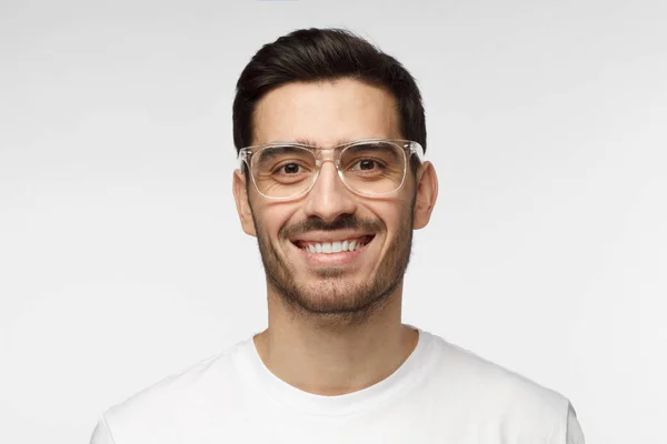 Closeup horizontal de homem branco bonito jovem com o cabelo do rosto isolado em fundo cinza vestido com camiseta branca e óculos de plástico transparente olhando para a câmera e sorrindo positivamente — Fotografia de Stock