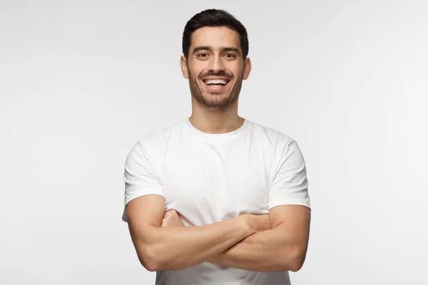 Retrato interior de medio cuerpo del joven caucásico europeo aislado sobre fondo gris de pie en camiseta blanca con los brazos cruzados, sonriendo y riendo positivamente mientras mira directamente a la cámara — Foto de Stock