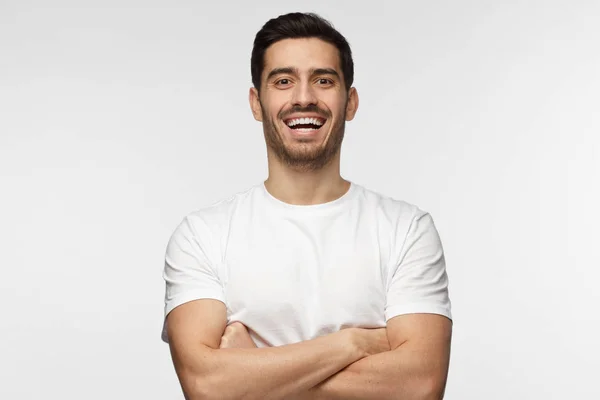 Horizontal closeup de jovem bonito europeu homem vestido de branco T-shirt retratado isolado no fundo cinza, seus braços cruzados, mostrando aberto amigável sorriso amigável feliz enquanto se comunica — Fotografia de Stock