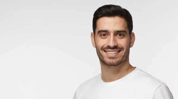 Primer plano de un joven caucásico europeo aislado sobre fondo gris usando una camiseta blanca casual con pelo corto oscuro y un poco de vello facial mostrando una sonrisa abierta feliz, espacio para copiar en el lado izquierdo — Foto de Stock