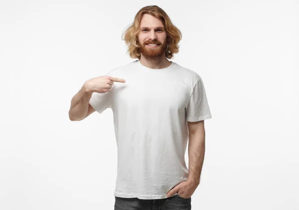 Half-length portrait of young handsome bearded male with long fair hair isolated on white background dressed in white blank T-shirt pointing to it with finger of right hand, copyspace for advertising — Stock Photo, Image