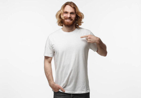 Indoor studio portrait of young Scandinavian man with face hair isolated on white background wearing white blank T-shirt pointing to it with finger of left hand, copyspace for advertising your company