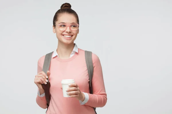 年轻的欧洲白人妇女的室内照片在灰色背景下被隔离戴着眼镜, 穿着随意和明亮的背包后面, 拿着外卖咖啡杯和微笑愉快 — 图库照片