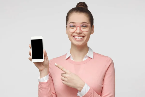 Imagem de close-up da jovem senhora europeia caucasiana retratada isolada no fundo cinza segurando celular em branco e mostrando-o com sorriso, espaço de cópia para publicidade de bens e serviços incluídos — Fotografia de Stock