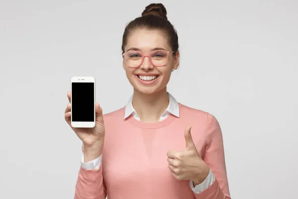 Retrato de estudio de una joven europea guapa vestida con jersey rosa y gafas, aislada sobre fondo gris que muestra la pantalla del teléfono inteligente en blanco con sonrisa, espacio para la publicidad — Foto de Stock