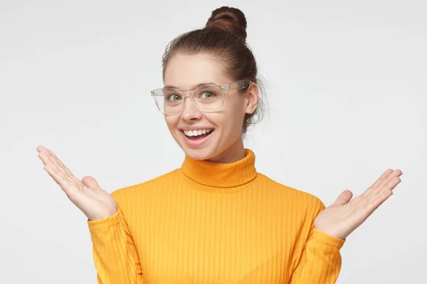 Retrato horizontal de una joven europea guapa en jersey de cuello alto amarillo y gafas aisladas sobre fondo gris sonriendo felizmente y haciendo gesto de la mano de agradable sorpresa y satisfacción — Foto de Stock