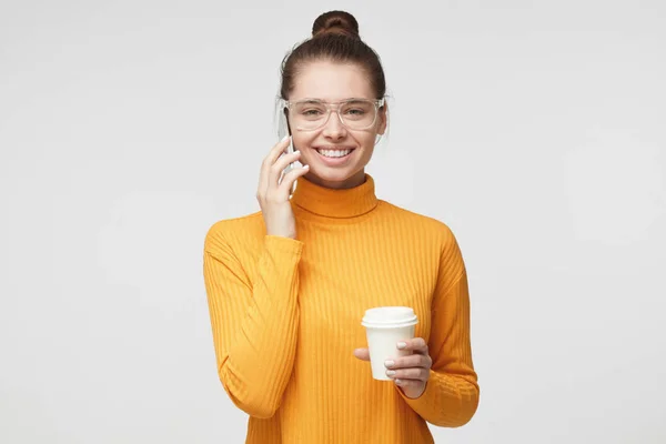 Foto close-up de jovem bonita fêmea europeia isolada em fundo cinza em pé em camisola amarela brilhante com copo de café de papelão, falando no celular com o companheiro, mostrando sorriso amigável feliz — Fotografia de Stock