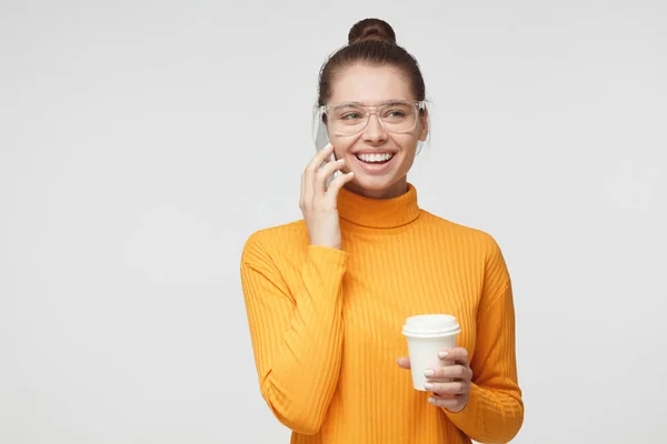 Retrato de close-up de jovem bela mulher retratada isolada em fundo cinza no topo amarelo, tendo óculos grandes, segurando xícara de papelão de café takeaway, sorrindo alegremente — Fotografia de Stock