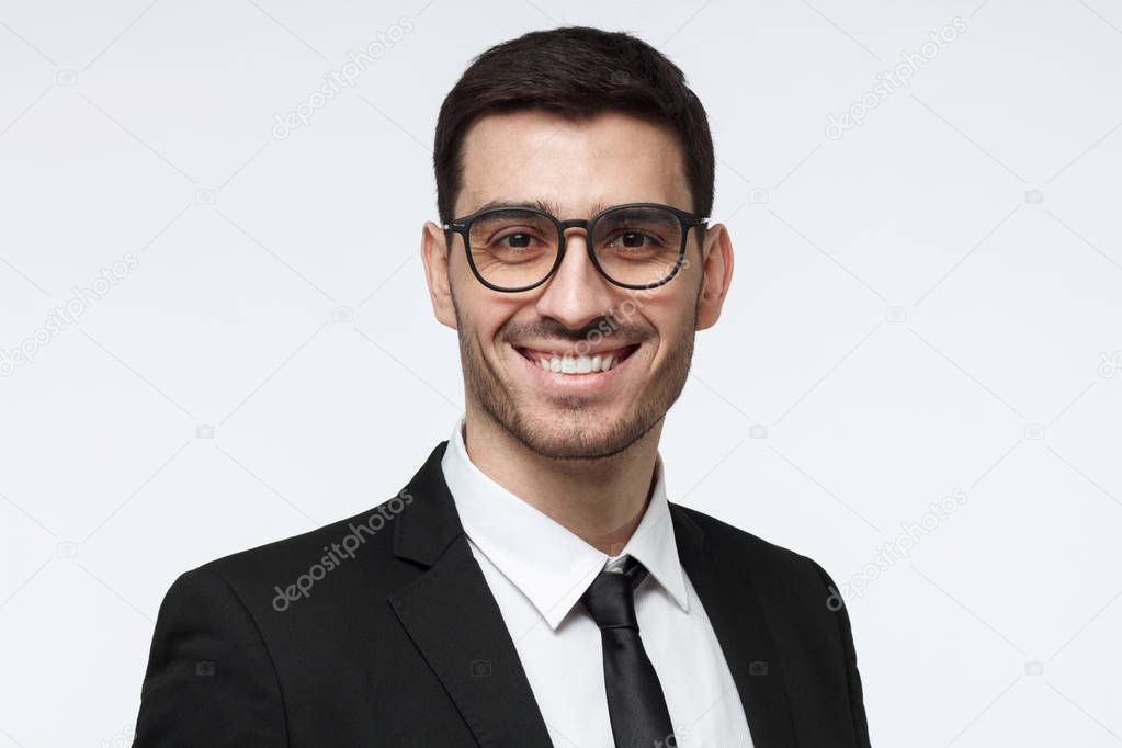 Horizontal headshot of young European Caucasian man in formal clothes isolated on gray background looking through glasses and smiling positively, feeling confident about success in his business