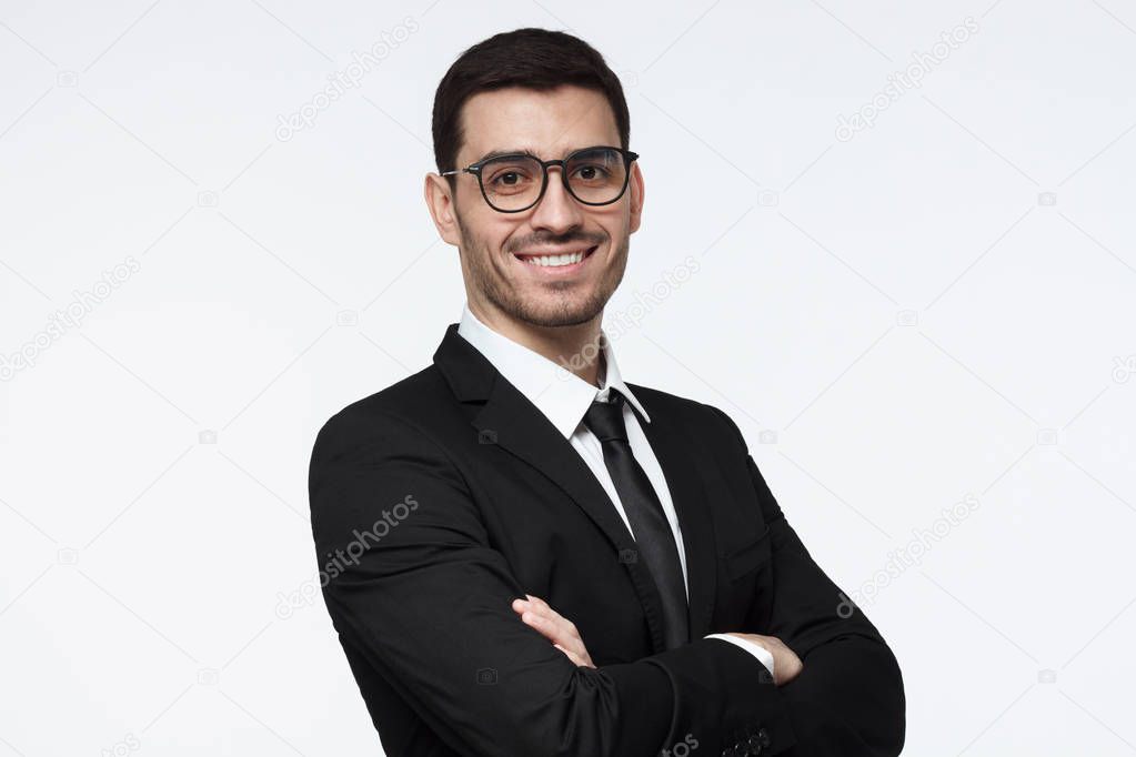 Horizontal photo of handsome Caucasian businessman isolated on white background wearing black suit, white shirt, tie and eyeglasses with arms crossed and positive face expression, ready to cooperate