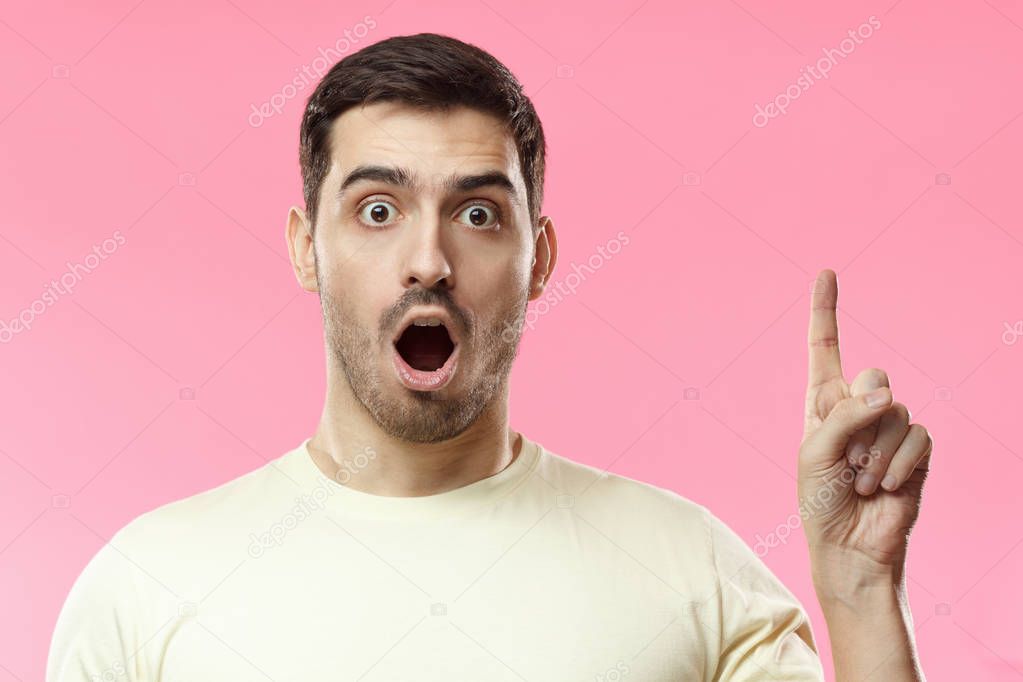 Indoor photo of young Caucasian guy isolated on pink background wearing white blank T-shirt having opened mouth in surprise and pointing upwards drawing attention to beneficial offer or sale