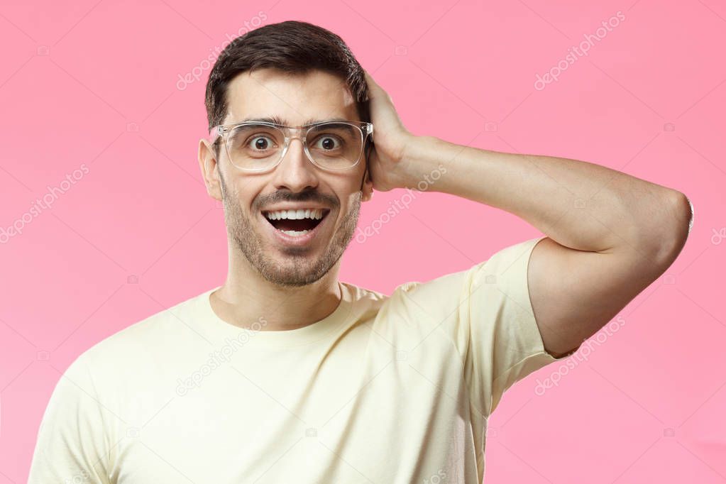 Horizontal closeup of young European guy isolated on pink background with beige casual T-shirt and transparent plastic glasses spreading folded arm and having opened eyes and mouth in deep surprise
