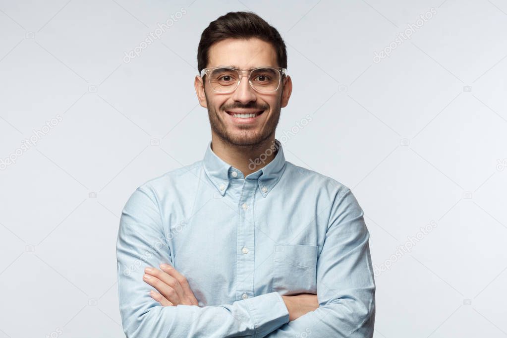Horizontal half-length portrait of handsome Caucasian man isolated on grey background standing with arms crossed and plastic safety eyeglasses on, showing positive smile to camera, looking confident