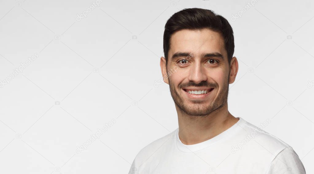 Closeup headshot of young European Caucasian man isolated on gray background wearing casual white T-shirt with short dark hair and some face hair showing happy open smile, copyspace in left side