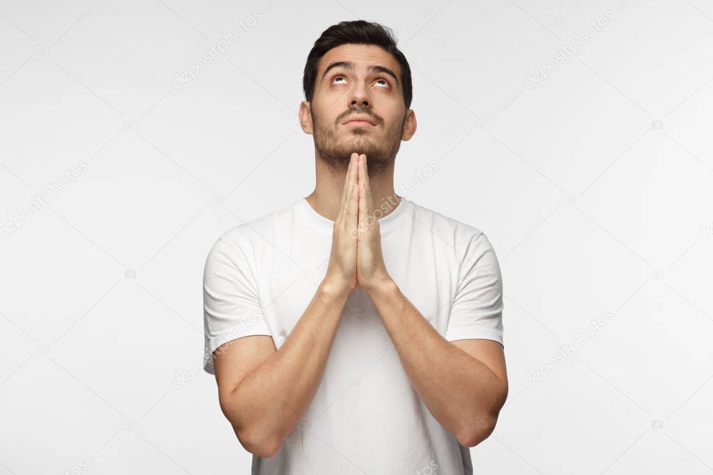 Indoor picture of young European Caucasian man pictured isolated on gray background dressed in white T-shirt praying with hands pressed together, looking upwards as if asking god for blessing