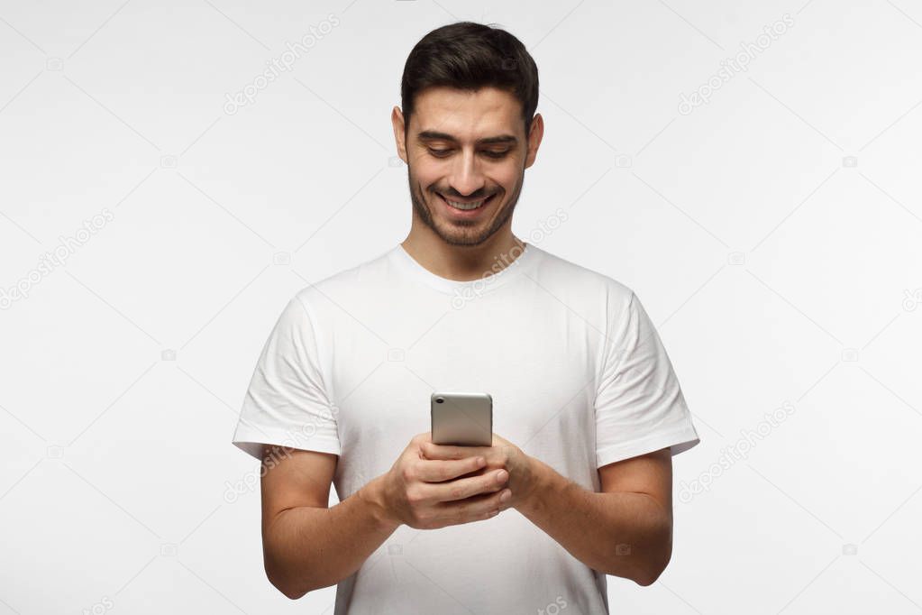 Horizontal photo of young good-looking European man pictured isolated on grey background dressed in casual blank T-shirt holding smartphone with bond hands and smiling positively while looking at it