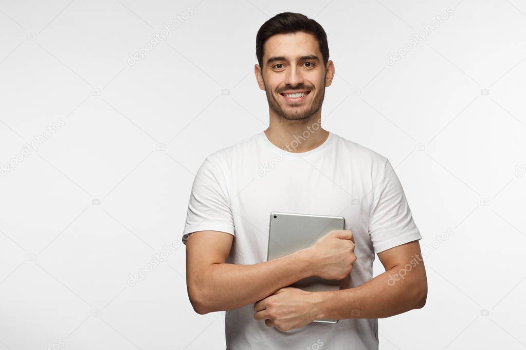 Horizontal portrait of young handsome European Caucasian man pictured isolated on grey background holding tablet computer next to his body, smiling, looking satisfied and ready to help and study