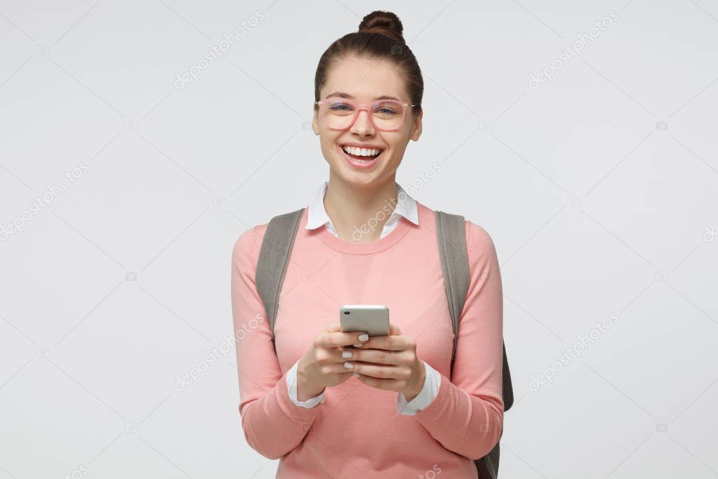Indoor portrait of young European Caucasian lady isolated on gray background wearing gray backpack and pink clothes and glasses, holding phone in hands, feeling happy and excited while reading texts