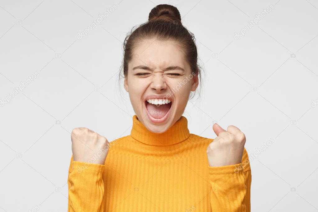 Closeup photo of young pretty European female isolated on grey background dressed in mustard turtleneck sweater screaming with open mouth and closed eyes, clenched fists as if feeling victory