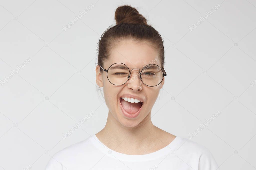 Closeup headshot of young good-looking teenage girl pictured isolated on gray background dressed in white T-shirt smiling and winking, having pulled tongue out as if joking or responding emotionally