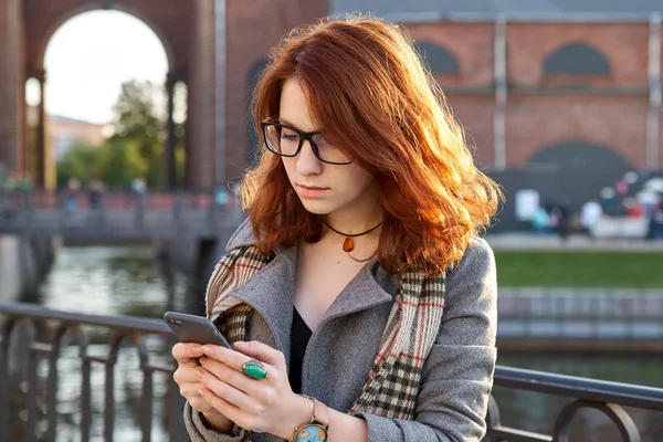 Modern vörös hajú fiatal nő használ szúró telefon őszi parkban. lány göndör gyömbér frizura, textil, mosolyogva, állandó kint esik. Élénk színek, természetes fény. — Stock Fotó