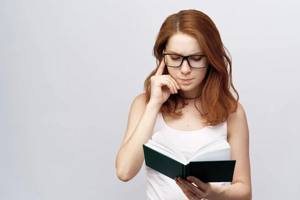 Ritratto di una bella ragazza dai capelli rossi con gli occhiali in un gilet che tiene un libro isolato su sfondo bianco. Una giovane donna attraente legge un libro concentrato, tiene il dito al tempio — Foto Stock