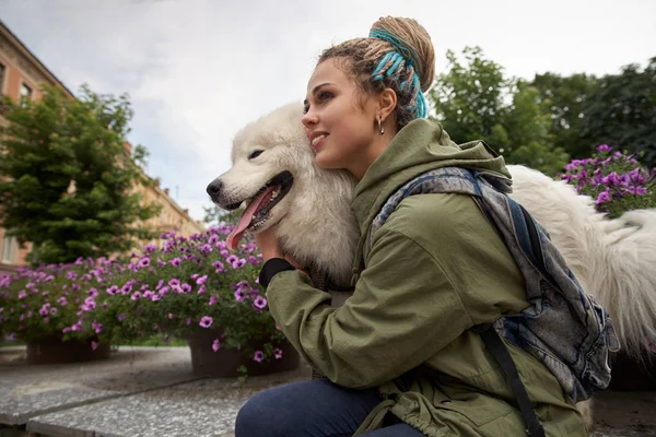 Cute young teen girl with dreadlocks on her head in a light green jacket and a backpack walks through a blooming Park with her four-legged friend Samoyed dog.
