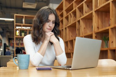 Kahve kupa ve dizüstü bilgisayar ile genç uzun saçlı beyaz tenli kız beyaz kazaklı kafede bir masada oturur