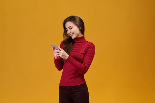 Porträtt av glada leende flicka isolerad på orange bakgrund, halv-vände kameran och hålla en telefon. — Stockfoto