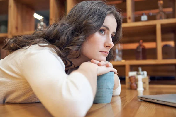 Attraente giovane ragazza dai capelli lunghi con una faccia annoiata piegata al tavolo in caffè e guarda attentamente in lontananza — Foto Stock