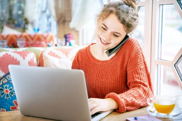 Όμορφη χαμογελαστή κοκκινομάλλα κοπέλα φακιδωμένος εργάζονται με το laptop στο εστιατόριο του πίνακα κατά τη διάρκεια μια συνομιλία στο τηλέφωνο. — Φωτογραφία Αρχείου