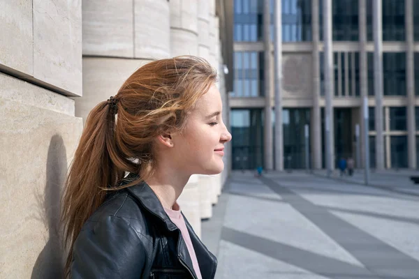 Retrato con el lado de la joven chica moderna en una chaqueta de cuero tomando el sol en el fondo de la arquitectura urbana moderna — Foto de Stock